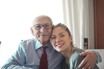 positive senior man in formal wear and eyeglasses hugging with young lady while sitting at table